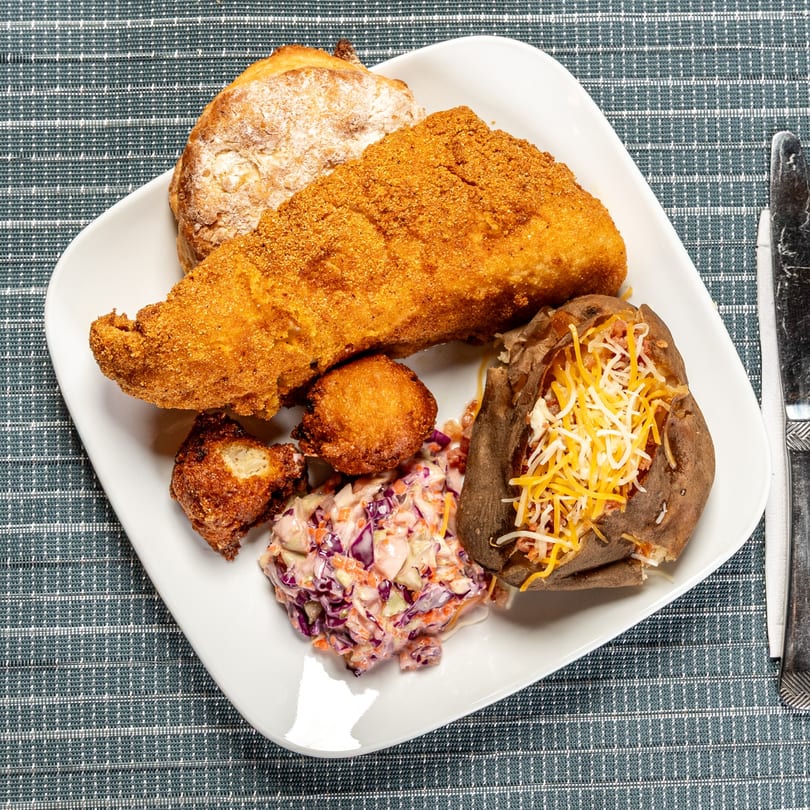 A fried fish platter with hush puppies, coleslaw, a baked potato with cheese, and a biscuit.