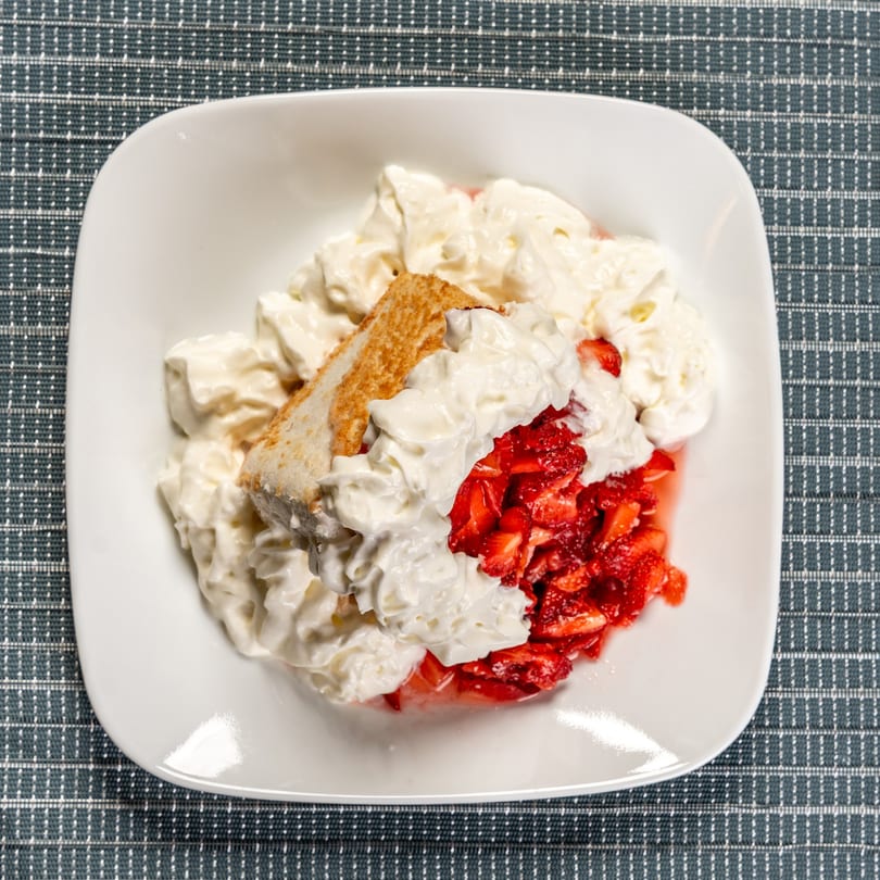 Strawberry shortcake with whipped cream and sliced strawberries in a white bowl on a blue placemat.