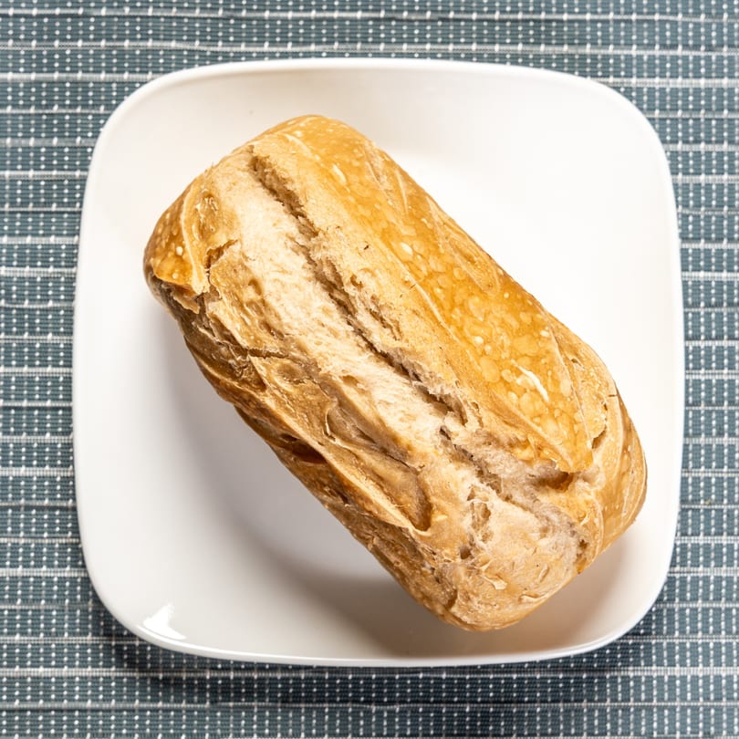 A golden-brown, crusty bread roll rests on a white square plate over a blue and white checkered tablecloth.
