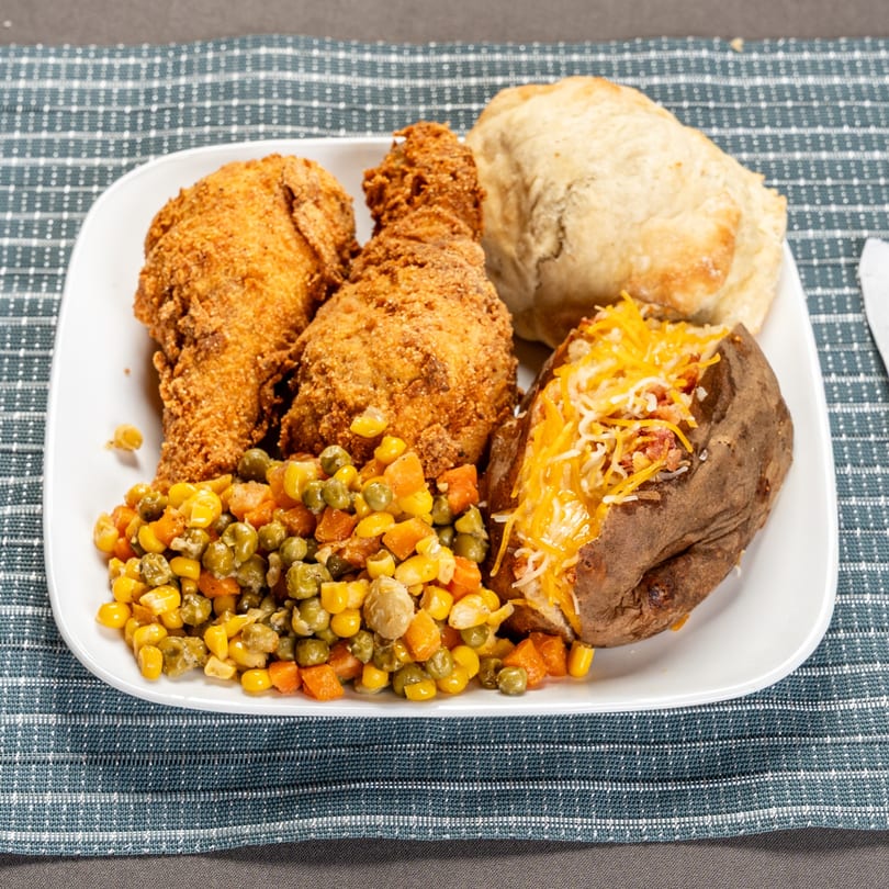 Fried chicken, mixed vegetables, a baked potato with cheese, and a biscuit on a white plate.